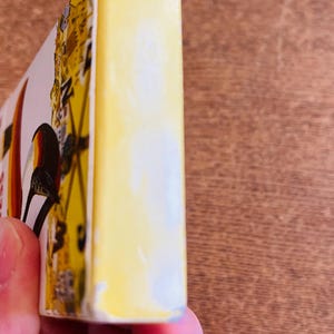May include: A yellow rectangular object with a bird illustration, held in a person's hand. The object has a white edge and a design with yellow, black, and red colors. The background is a wooden surface.