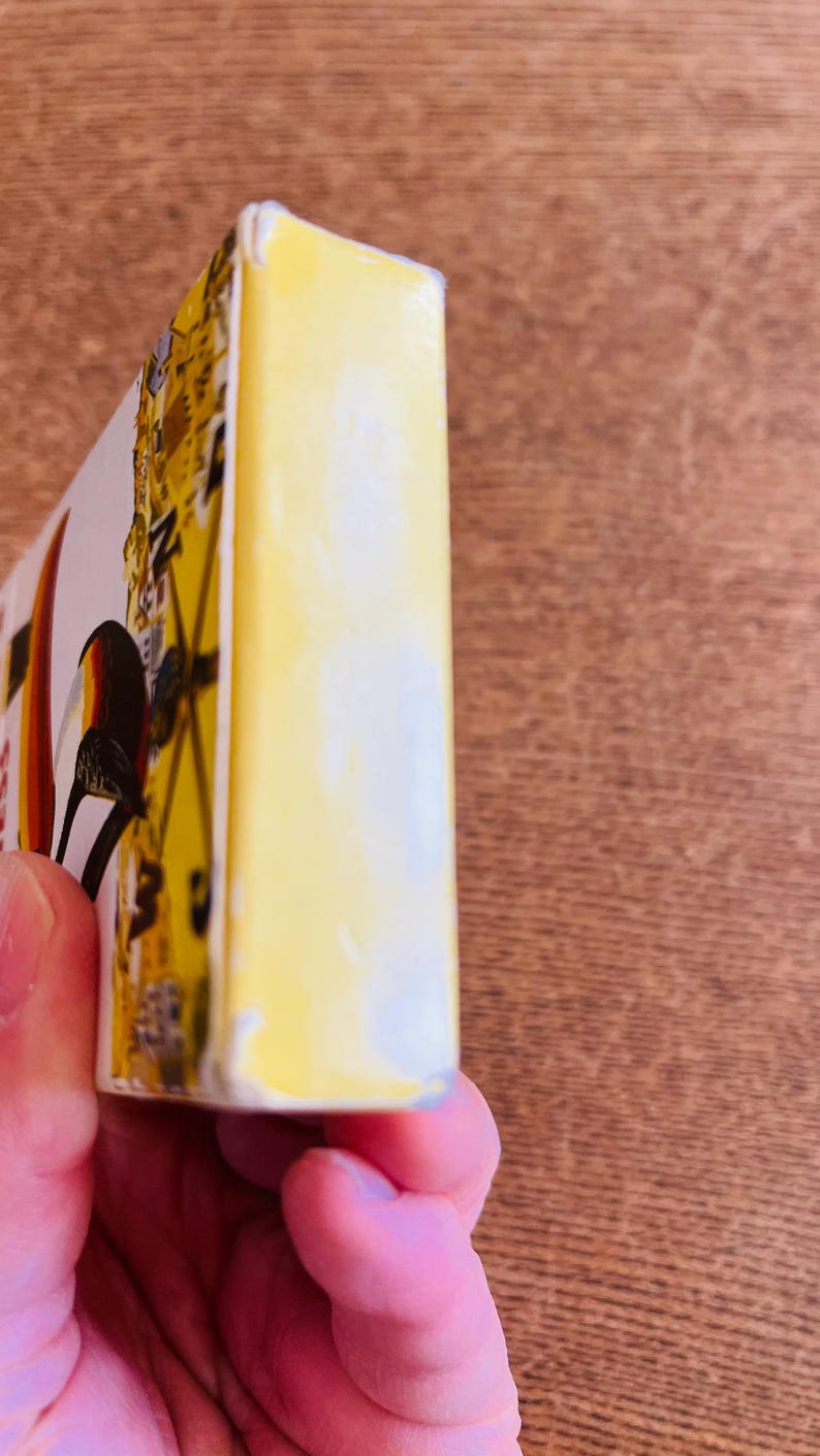 May include: A yellow rectangular object with a bird illustration, held in a person's hand. The object has a white edge and a design with yellow, black, and red colors. The background is a wooden surface.