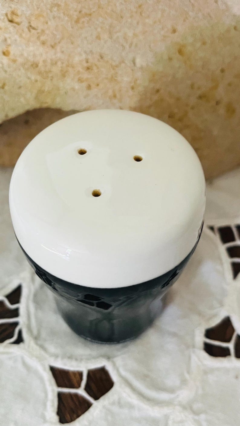 Op de afbeelding: Een witte en zwarte keramische zoutstrooier met drie gaatjes bovenop. De zoutstrooier heeft een glanzende afwerking en een cilindrische vorm. De basis is zwart en de bovenkant is wit. De zoutstrooier staat op een wit oppervlak.
