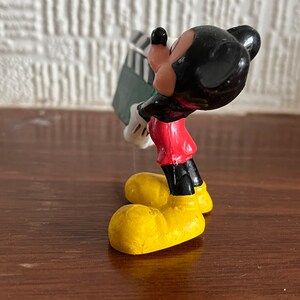 May include: A small plastic figurine of Mickey Mouse holding a green and black clapperboard. Mickey is wearing his signature red shorts, yellow shoes, and black gloves.