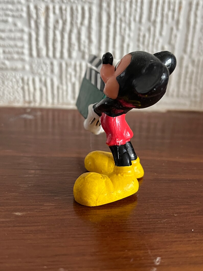May include: A small plastic figurine of Mickey Mouse holding a green and black clapperboard. Mickey is wearing his signature red shorts, yellow shoes, and black gloves.