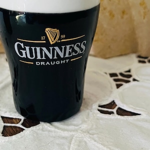 Op de afbeelding: Een zwart Guinness-bierglas met een witte bovenkant. Het Guinness-logo is wit met gouden accenten. Het glas staat op een wit kanten tafelkleed.