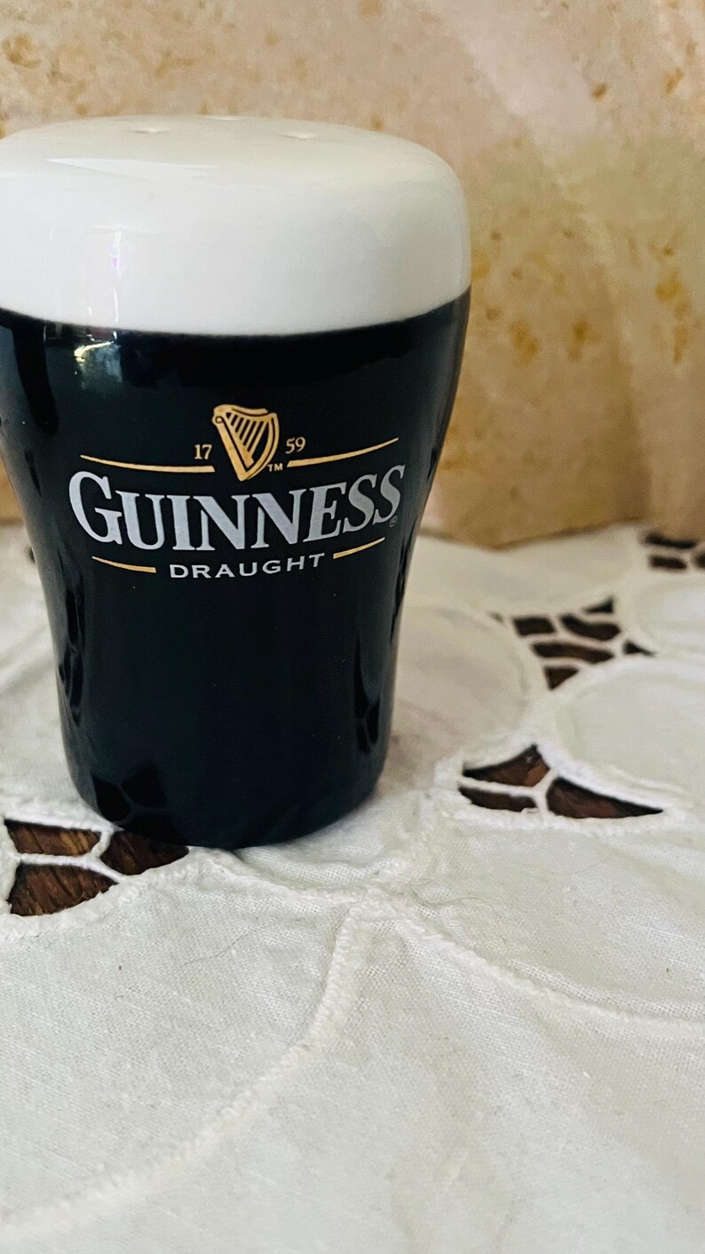 Op de afbeelding: Een zwart Guinness-bierglas met een witte bovenkant. Het Guinness-logo is wit met gouden accenten. Het glas staat op een wit kanten tafelkleed.