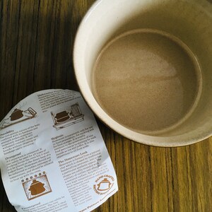 May include: A close-up of a beige stoneware bowl with a brown rim. The bowl is sitting on a wooden surface. A folded piece of paper with text about the stoneware is in the foreground. The text includes information about the durability, ovenproof, freezerproof, and hygienic qualities of the stoneware. The text also mentions that the stoneware is handcrafted in Britain.