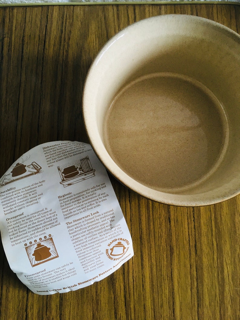 May include: A close-up of a beige stoneware bowl with a brown rim. The bowl is sitting on a wooden surface. A folded piece of paper with text about the stoneware is in the foreground. The text includes information about the durability, ovenproof, freezerproof, and hygienic qualities of the stoneware. The text also mentions that the stoneware is handcrafted in Britain.