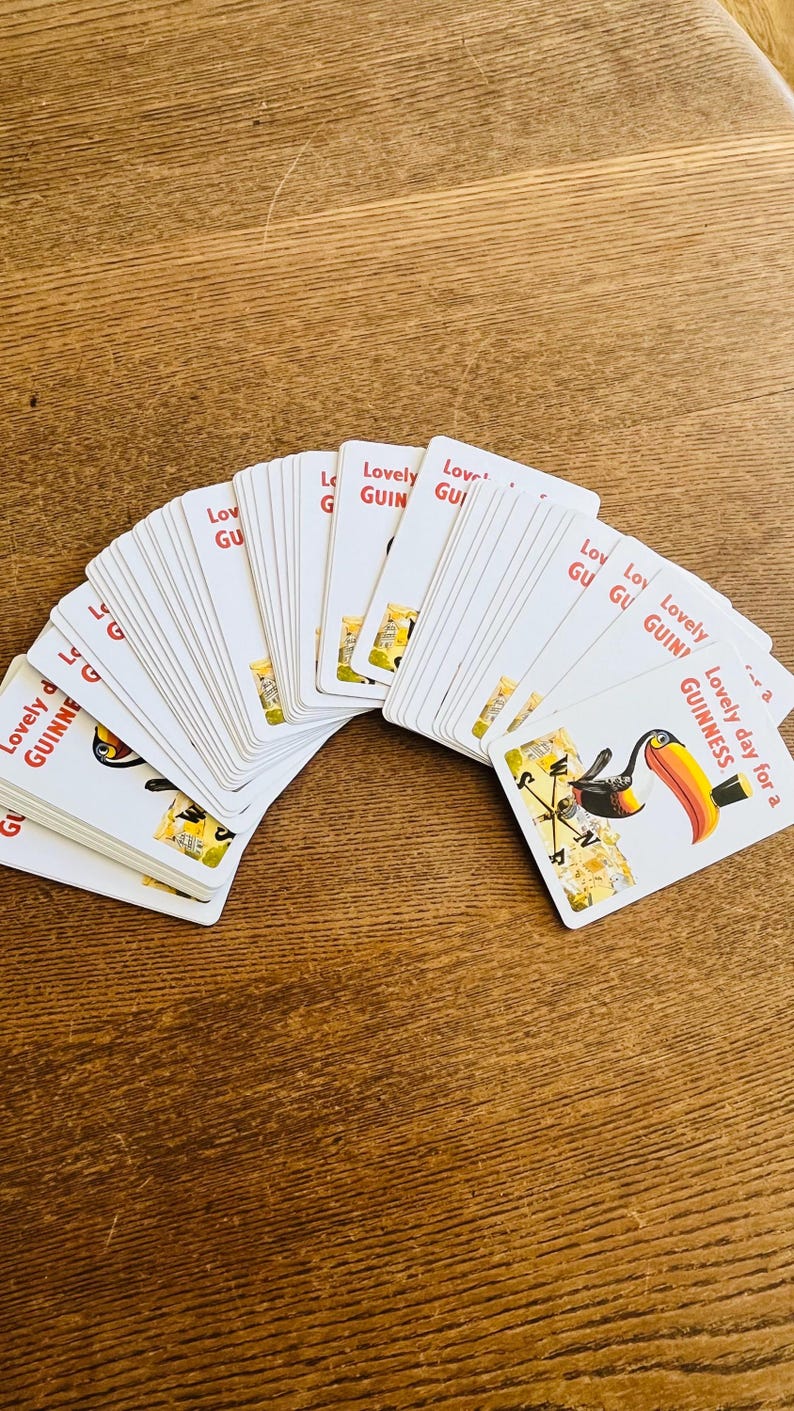 May include: A deck of white playing cards with the text "Lovely Guinness" and a toucan illustration. The cards are fanned out on a wooden surface, showcasing the design.