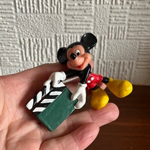 May include: A black and red plastic Mickey Mouse figurine holding a green and white clapperboard.