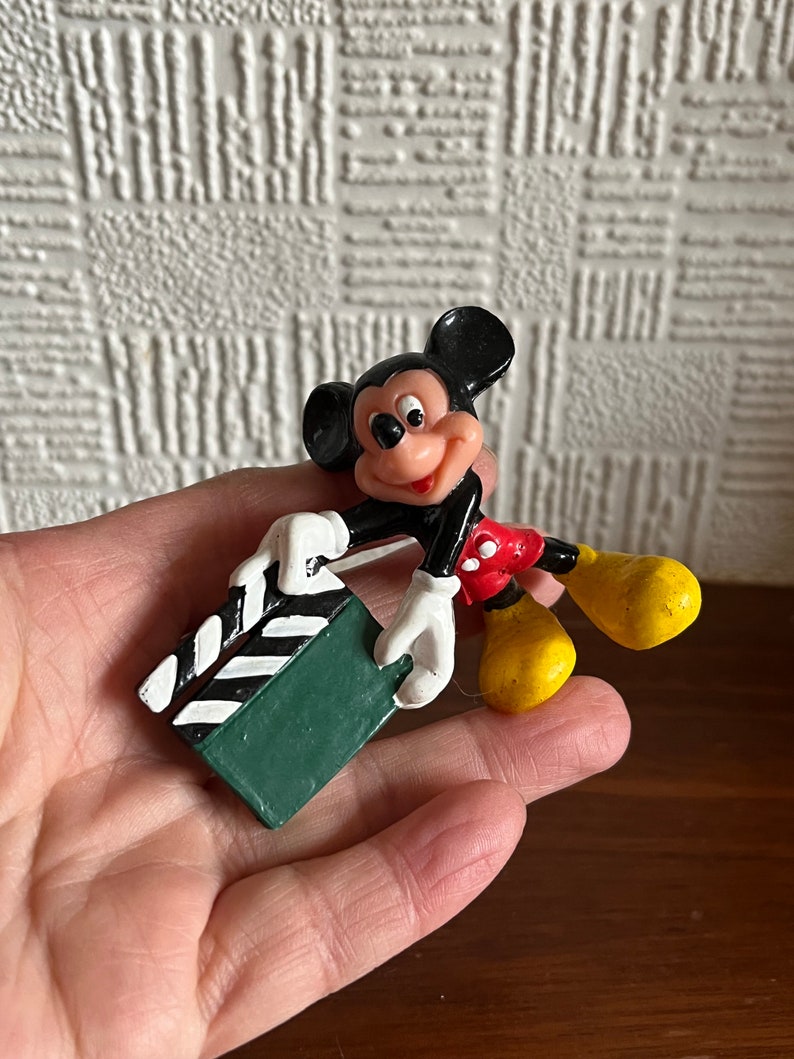 May include: A black and red plastic Mickey Mouse figurine holding a green and white clapperboard.