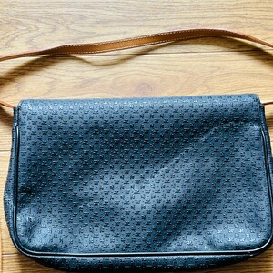 May include: A black shoulder bag with a repeating pattern across the front. The bag has a brown leather strap and is set against a wooden background.