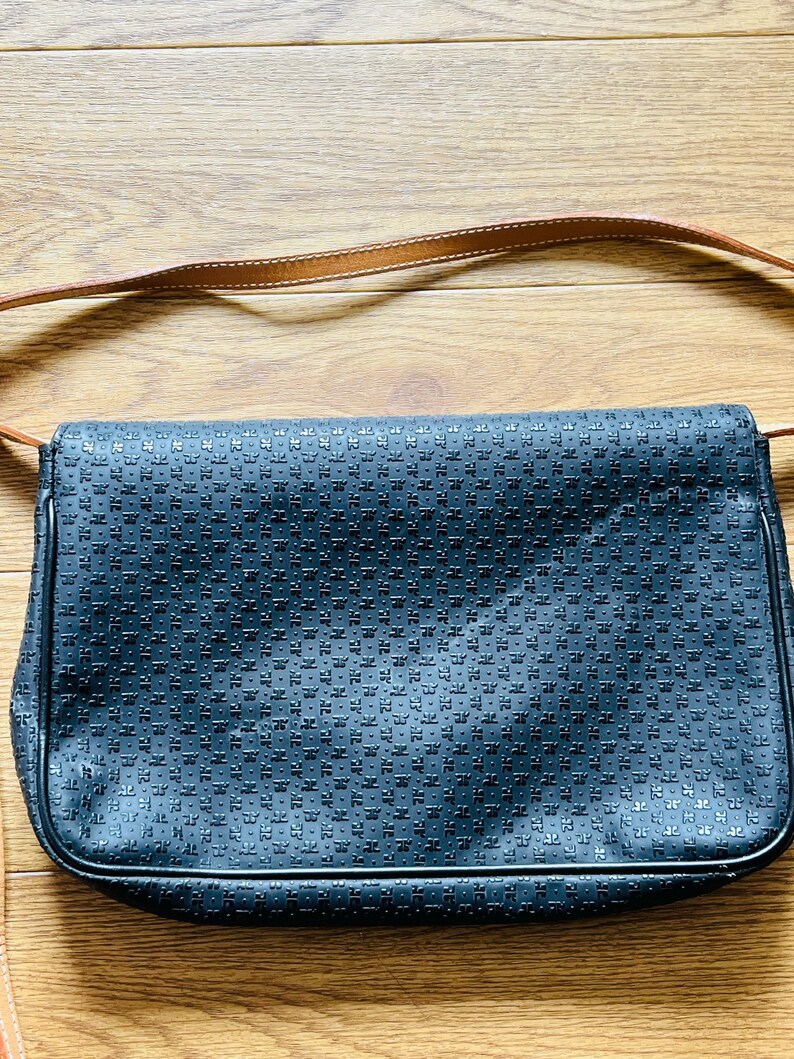 May include: A black shoulder bag with a repeating pattern across the front. The bag has a brown leather strap and is set against a wooden background.