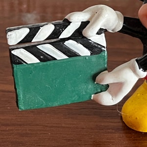 May include: A green and black clapperboard held by a Mickey Mouse figurine. The figurine is made of painted plastic and has a yellow body, white gloves, and black shoes.