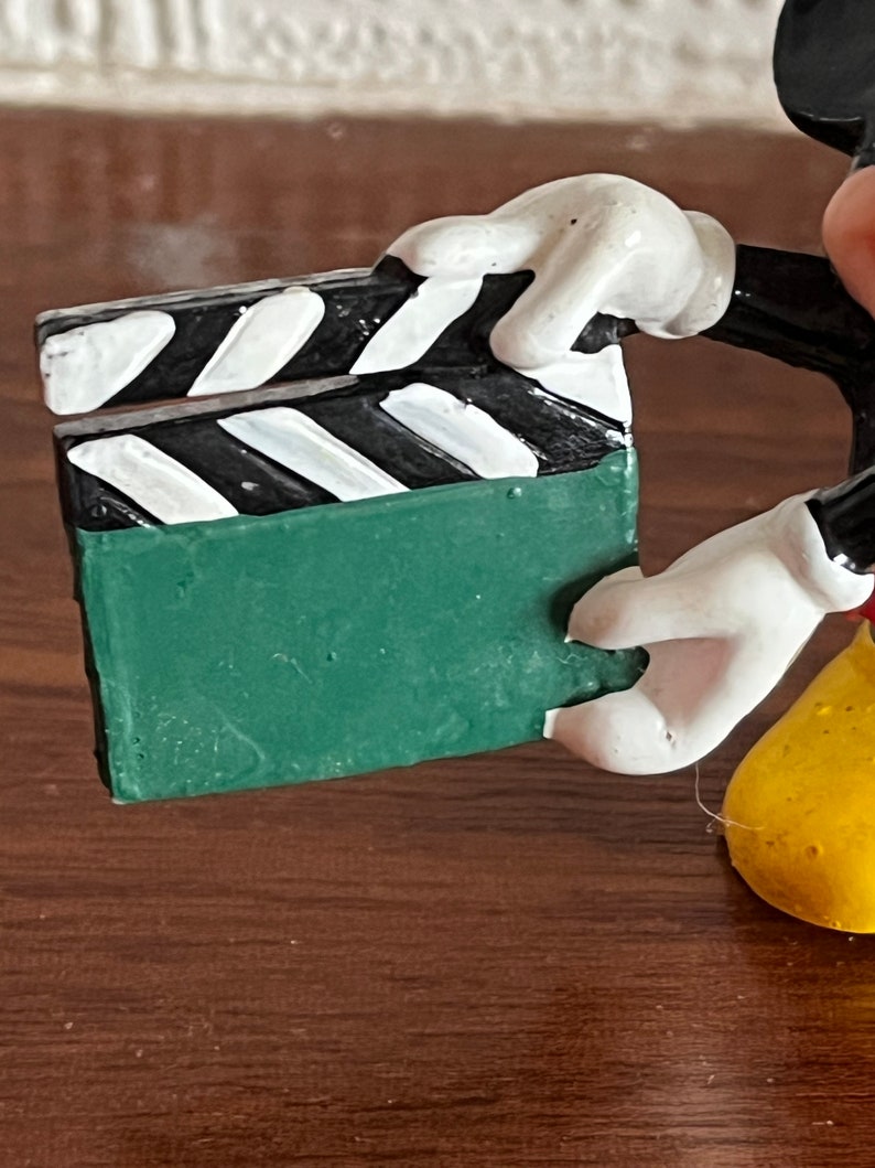 May include: A green and black clapperboard held by a Mickey Mouse figurine. The figurine is made of painted plastic and has a yellow body, white gloves, and black shoes.