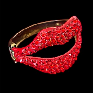 May include: A red rhinestone-covered bracelet with a gold-tone metal base. The bracelet is a hinged style, with a curved design. The red rhinestones are densely packed, creating a sparkling effect. The background is black.