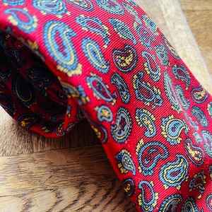 May include: A red paisley tie featuring a repeating pattern of blue and yellow teardrop-shaped designs. The tie is made of a smooth material and is displayed on a wooden surface. A classic accessory for formal or smart casual wear.