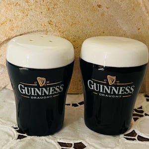 Op de afbeelding: Een paar keramische zout- en peperstrooiers in de vorm van Guinness pintglazen. De strooiers zijn zwart met witte bovenkanten en hebben het Guinness-logo in goud. Het logo bevat het woord "Guinness" en het woord "Draught".