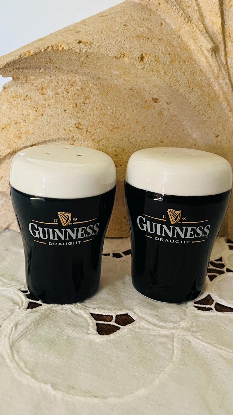 Op de afbeelding: Een paar keramische zout- en peperstrooiers in de vorm van Guinness pintglazen. De strooiers zijn zwart met witte bovenkanten en hebben het Guinness-logo in goud. Het logo bevat het woord "Guinness" en het woord "Draught".