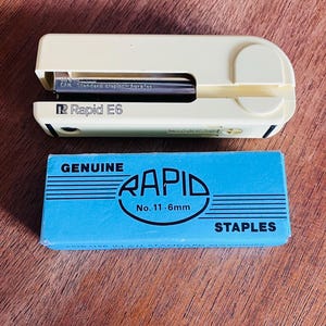 May include: A cream-colored Rapid E6 stapler with a black staple cartridge, and a blue box of Rapid staples. The box has the word "GENUINE" above the Rapid logo and "STAPLES" below. The stapler is on a wooden surface.