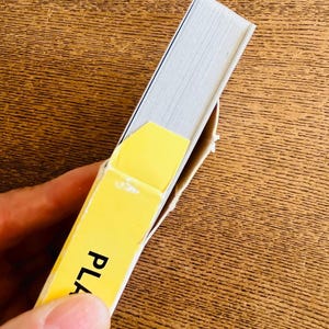 May include: A partially visible book with white pages and a yellow cover. The word "PLA" is printed on the yellow cover. The book is held by a person.