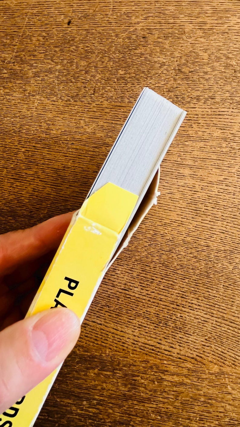 May include: A partially visible book with white pages and a yellow cover. The word "PLA" is printed on the yellow cover. The book is held by a person.