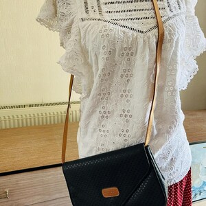 May include: Black crossbody bag with a perforated design and a tan leather strap. The bag has a rectangular shape with a flap closure and a small tan leather patch. The bag is hanging on a white eyelet blouse.