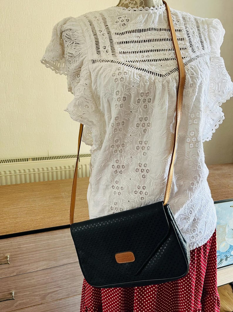 May include: Black crossbody bag with a perforated design and a tan leather strap. The bag has a rectangular shape with a flap closure and a small tan leather patch. The bag is hanging on a white eyelet blouse.