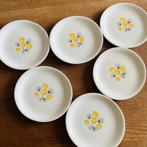 以下が含まれることがあります： 黄色とグレーの花柄が描かれた、白いセラミックプレートが6枚。木製の表面に配置されています。各プレートの中央に花の模様があります。