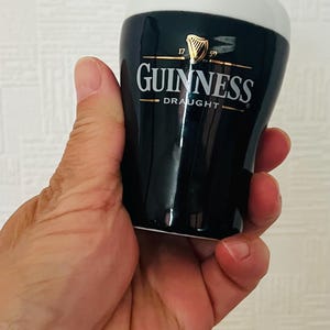 Op de afbeelding: Een zwart en wit Guinness draught keramisch shotglaasje. Het glas heeft het Guinness-logo in gouden letters en een witte bovenkant. Het shotglaasje wordt in een hand gehouden.