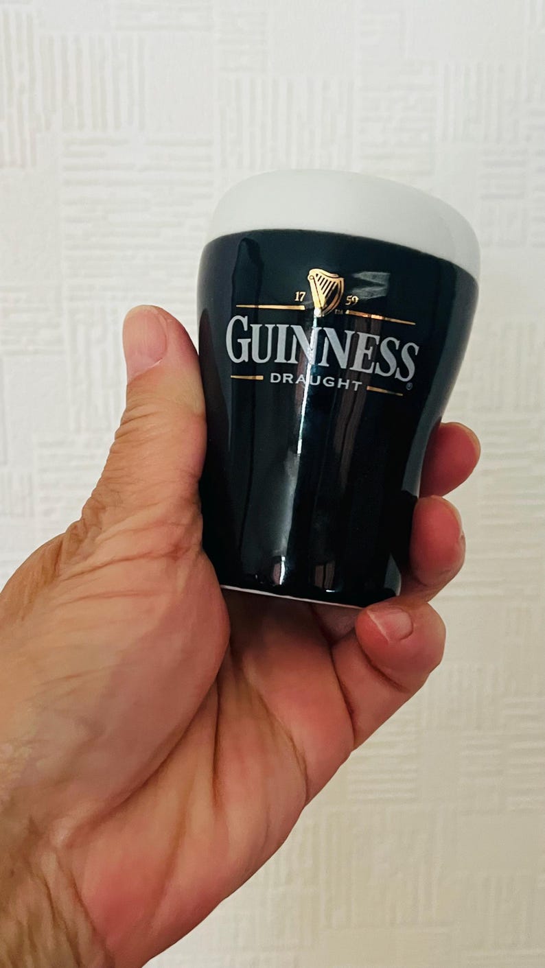 Op de afbeelding: Een zwart en wit Guinness draught keramisch shotglaasje. Het glas heeft het Guinness-logo in gouden letters en een witte bovenkant. Het shotglaasje wordt in een hand gehouden.