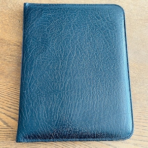 May include: A dark teal leather-bound notebook or journal. The cover has a textured, pebbled appearance. The notebook is rectangular with rounded corners and appears to be closed. The background is a light brown wooden surface.