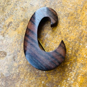 May include: A handcrafted wooden brooch in a curved, comma-like shape. The brooch is made of dark brown wood with visible grain patterns and a brass pin backing. The design is simple and elegant, suitable for various outfits.