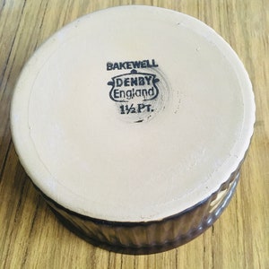 May include: A brown ceramic baking dish with a white interior. The bottom of the dish has a black stamp that reads "Bakewell Denby England 1 1/2 Pt."