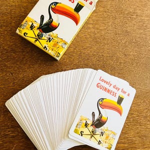 May include: A deck of Guinness playing cards with a toucan illustration and the text "Lovely day for a Guinness." The cards are white with a yellow border and are fanned out on a wooden surface. A matching card box is also present.