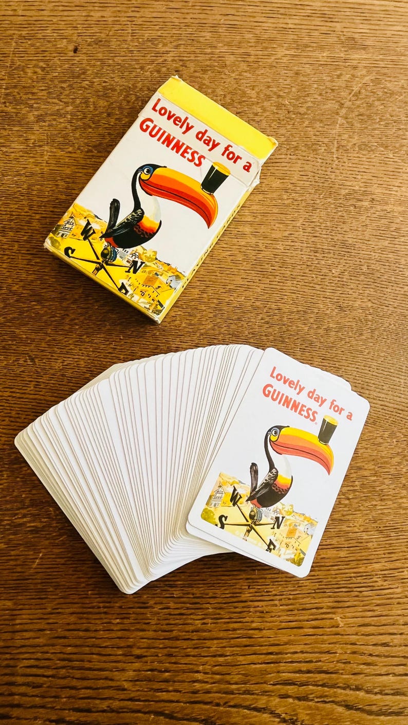 May include: A deck of Guinness playing cards with a toucan illustration and the text "Lovely day for a Guinness." The cards are white with a yellow border and are fanned out on a wooden surface. A matching card box is also present.