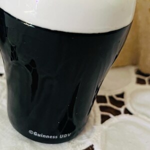 Op de afbeelding: Een keramische zoutstrooier in de vorm van een Guinness pintglas. De bovenkant is wit en de onderkant is zwart. De Guinness- en UDV-logo's zijn op de zijkant gedrukt. De strooier heeft twee gaten aan de bovenkant.
