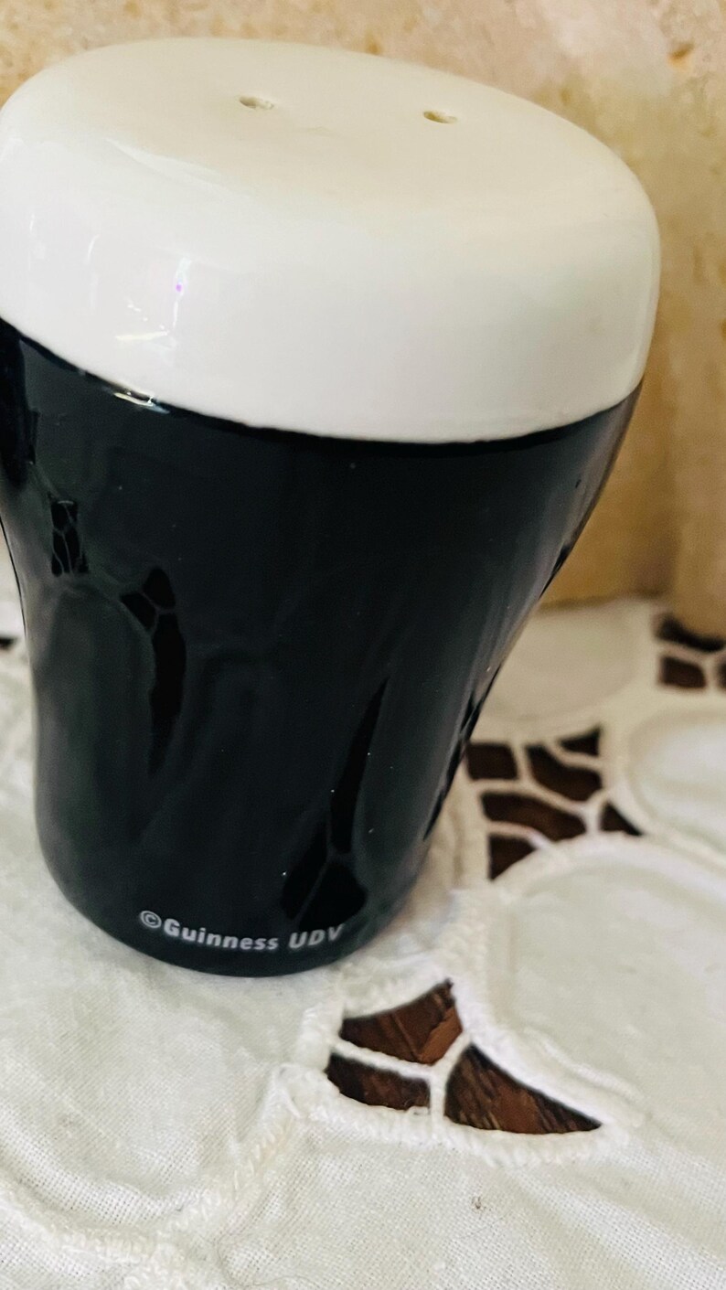 Op de afbeelding: Een keramische zoutstrooier in de vorm van een Guinness pintglas. De bovenkant is wit en de onderkant is zwart. De Guinness- en UDV-logo's zijn op de zijkant gedrukt. De strooier heeft twee gaten aan de bovenkant.
