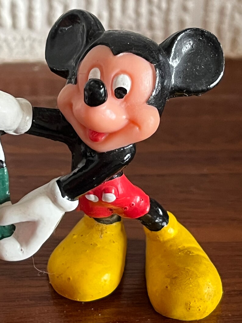 May include: A black and red plastic figurine of Mickey Mouse. Mickey is wearing yellow shoes and has a big smile.