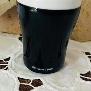 Op de afbeelding: Een keramische zoutstrooier in de vorm van een Guinness stout pint. De bovenkant is wit en de body zwart, met het Guinness UDV-logo op de basis. Het staat op een wit kanten tafelkleed.