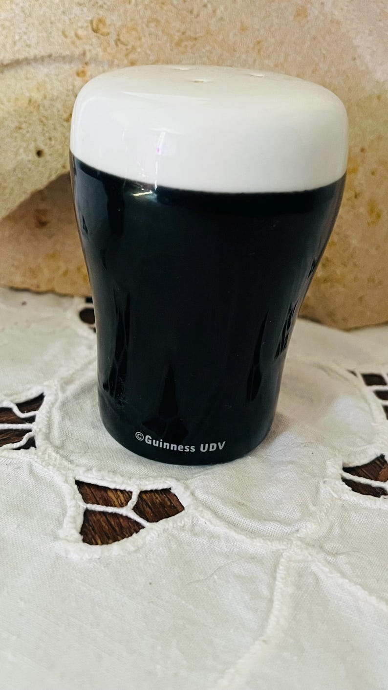 Op de afbeelding: Een keramische zoutstrooier in de vorm van een Guinness stout pint. De bovenkant is wit en de body zwart, met het Guinness UDV-logo op de basis. Het staat op een wit kanten tafelkleed.