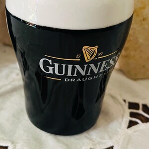 Op de afbeelding: Een keramische Guinness draught zout- en peperstrooier set. De bovenkant is wit en de onderkant zwart met het Guinness logo en het harpsymbool. De shaker staat op een witte, bedrukte doek.