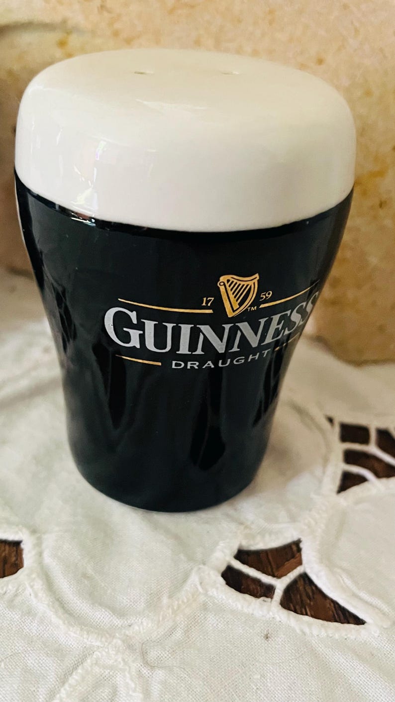 Op de afbeelding: Een keramische Guinness draught zout- en peperstrooier set. De bovenkant is wit en de onderkant zwart met het Guinness logo en het harpsymbool. De shaker staat op een witte, bedrukte doek.