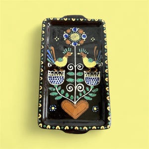 May include: Rectangular black ceramic tray with a colourful folk art design. The tray features two yellow birds, a brown heart, and floral elements in blue, green, and brown. The border is decorated with small yellow and blue dots.
