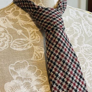 May include: A houndstooth patterned tie, with a repeating pattern of small black, red, and white squares. The tie is knotted and displayed on a mannequin, highlighting the fabric's texture and colour variations.