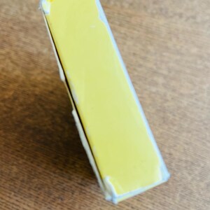 May include: A rectangular, yellow box with white edges, slightly angled. The box is on a wooden surface. The box appears to be sealed or wrapped in clear plastic.
