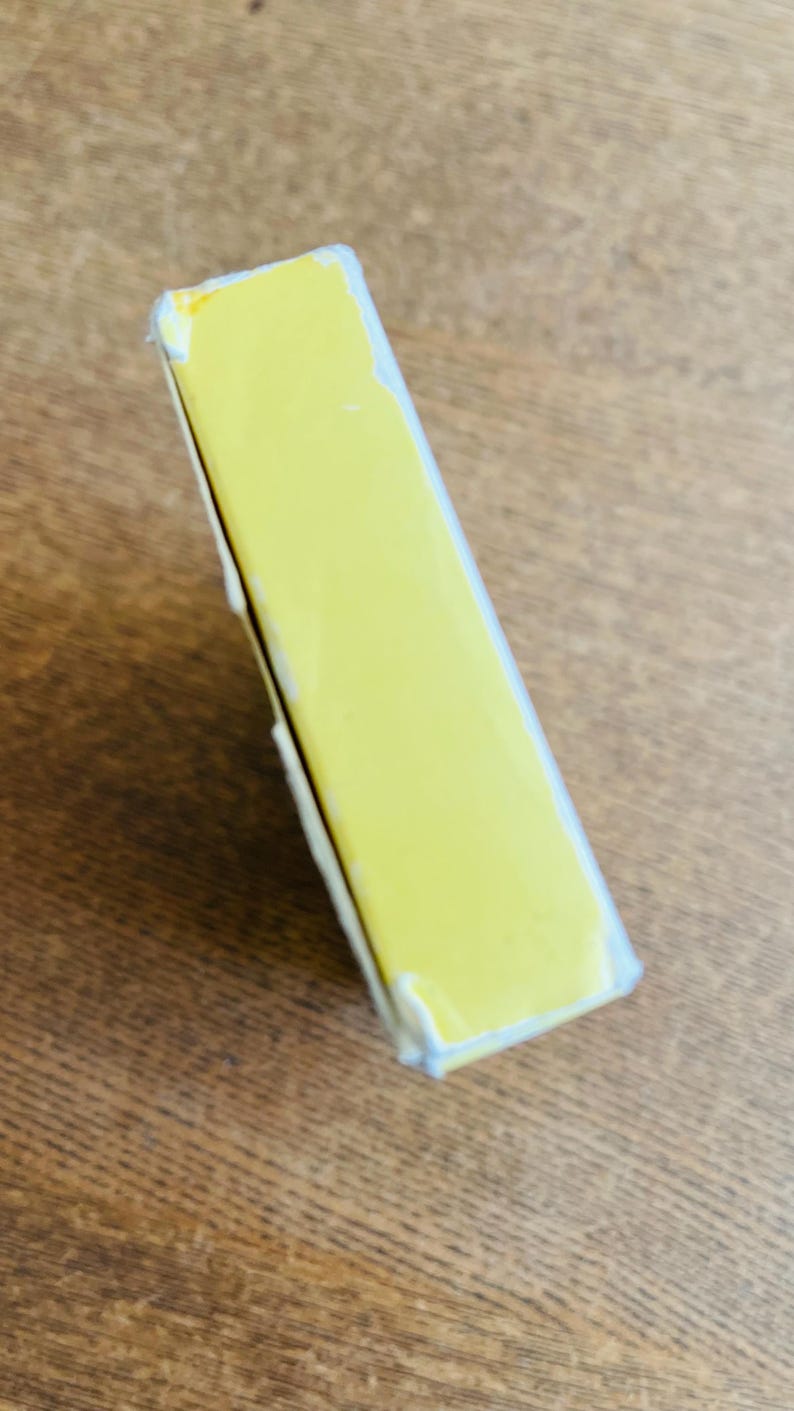 May include: A rectangular, yellow box with white edges, slightly angled. The box is on a wooden surface. The box appears to be sealed or wrapped in clear plastic.