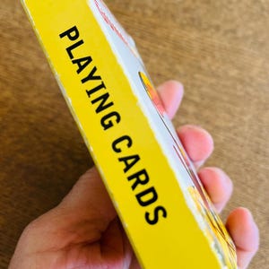 May include: A yellow playing cards box with the words "PLAYING CARDS" in bold black letters. The box is held in a person's hand. The box has a white and red design on the side.