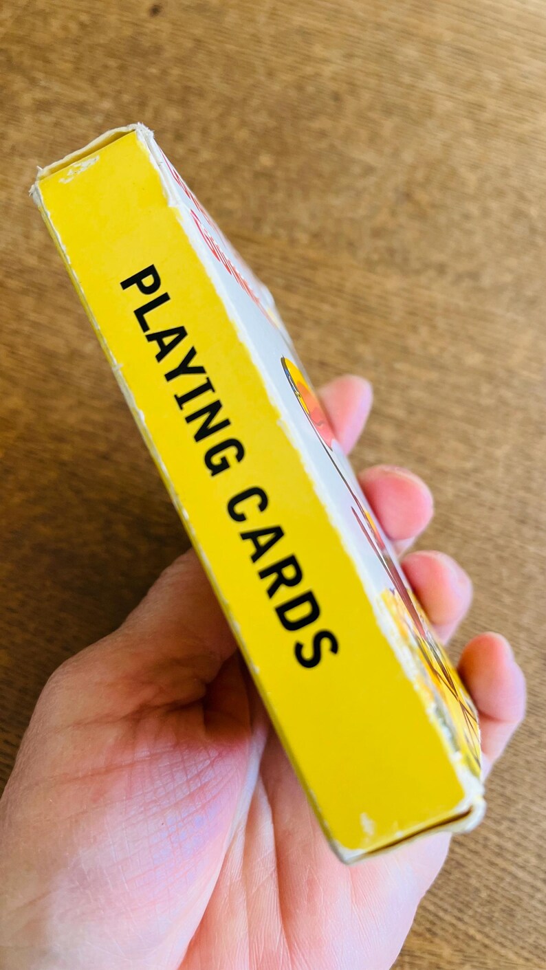 May include: A yellow playing cards box with the words "PLAYING CARDS" in bold black letters. The box is held in a person's hand. The box has a white and red design on the side.