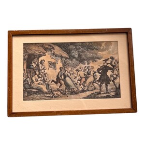 May include: Framed antique print depicting a lively outdoor scene with figures dancing and a musician playing. The artwork, in shades of gray and brown, is set in a wooden frame with a cream-colored matting, showcasing a vintage aesthetic.