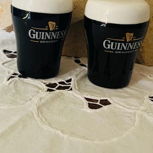 Op de afbeelding: Twee keramische Guinness-bierglazen met witte schuimtoppen. De zwarte glazen hebben het Guinness-logo en de tekst. De glazen staan op een wit kanten tafelkleed en een beige stenen achtergrond.