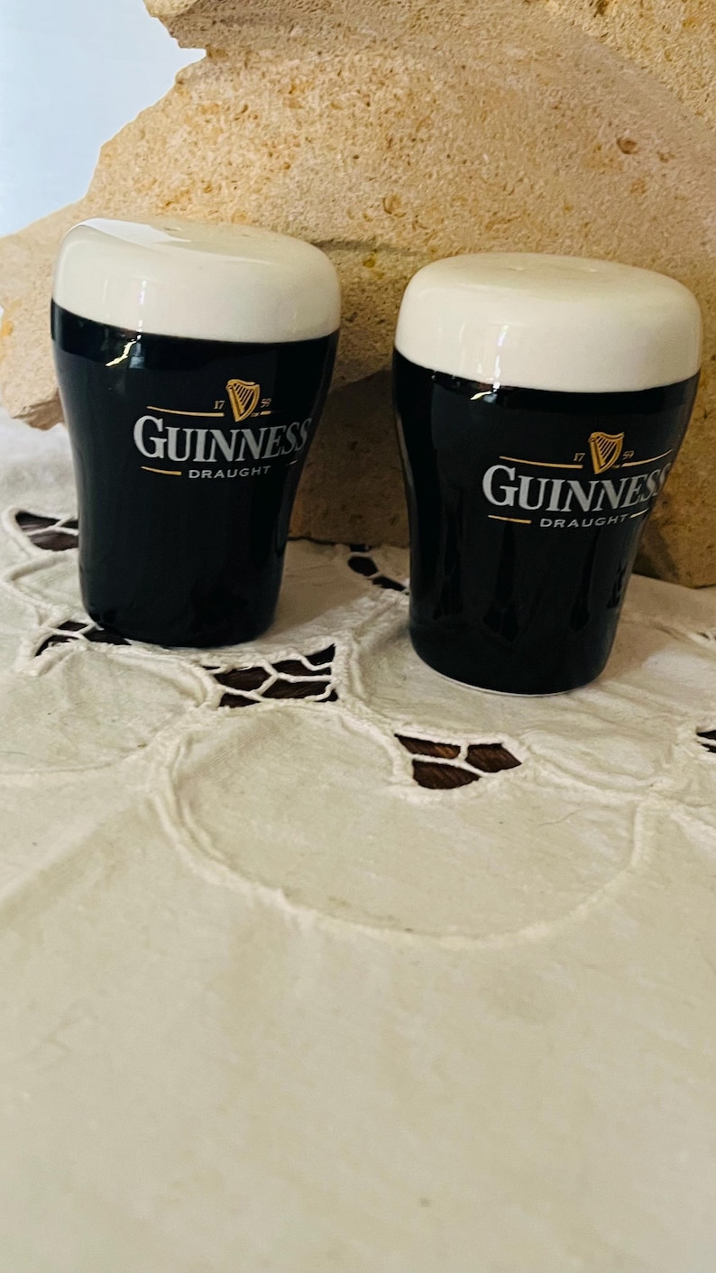 Op de afbeelding: Twee keramische Guinness-bierglazen met witte schuimtoppen. De zwarte glazen hebben het Guinness-logo en de tekst. De glazen staan op een wit kanten tafelkleed en een beige stenen achtergrond.