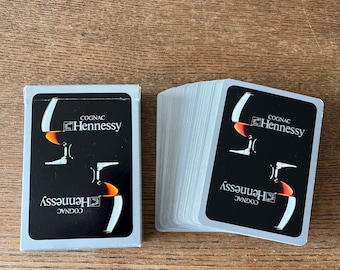 May include: A deck of playing cards with a black and white design featuring the Hennessy Cognac logo. The logo includes a glass of cognac with a flame inside. The cards are in a white box with the Hennessy Cognac logo on the front.